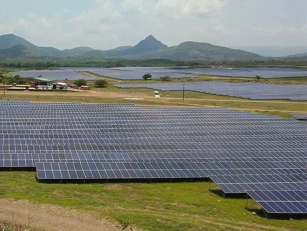 A large solar panel farm with mountains in the background under a cloudy sky.