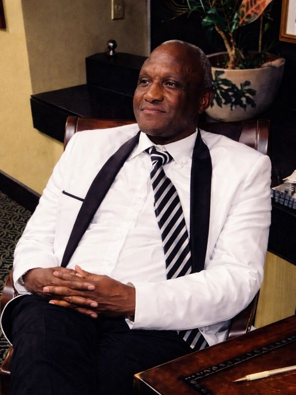 Man in white tuxedo with black lapels sitting confidently in a chair indoors.